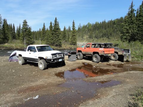 1990 Chevrolet Suburban - Two off-road vehicles are parked near a river, with a tent set up and a dense forest behind them.