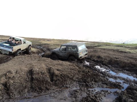 1990 Chevrolet Suburban - Two off-road vehicles navigate a challenging muddy terrain, with one truck carrying passengers and the other appearing stuck.