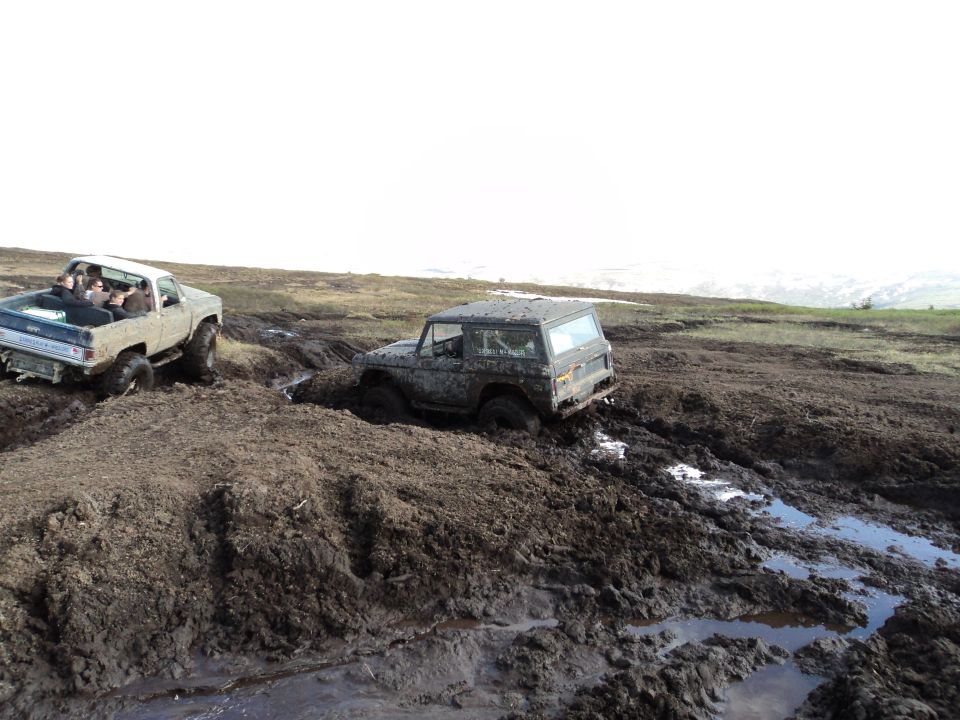 Two muddy off-road vehicles stuck in mud, one a pickup truck with passengers, the other a dark SUV