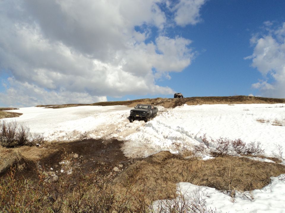 Two off-road vehicles navigate a snowy, hilly terrain under a cloudy blue sky