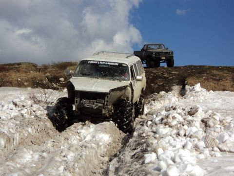 1990 Chevrolet Suburban - Two off-road vehicles navigate a snowy and muddy terrain under a cloudy sky.