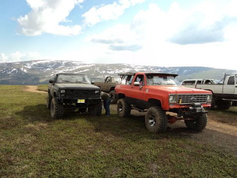 1990 Chevrolet Suburban - Two lifted SUVs, an orange Chevrolet Suburban and a black SUV, are parked on a grassy hill with snow-capped mountains in the background.