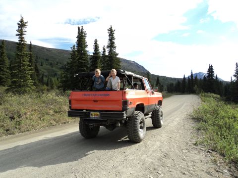 1990 Chevrolet Suburban - Lifted orange pickup truck with children in the bed travels a dirt road through a forested mountain landscape.