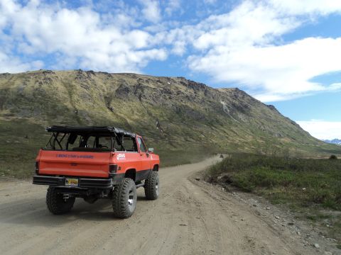 1990 Chevrolet Suburban - An orange lifted SUV with a roof rack travels down a dusty dirt road, with a mountain range in the background.