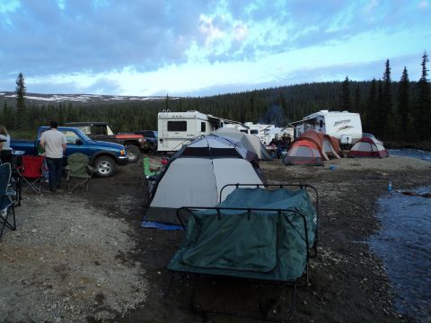1990 Chevrolet Suburban - A busy campsite is set up along a riverbank, with several tents and recreational vehicles under a cloudy sky.