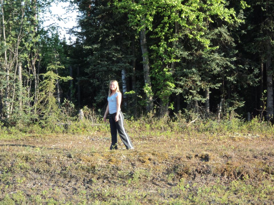Woman in light blue shirt and dark pants stands in a clearing with trees behind her. Supermotors.net logo in lower right