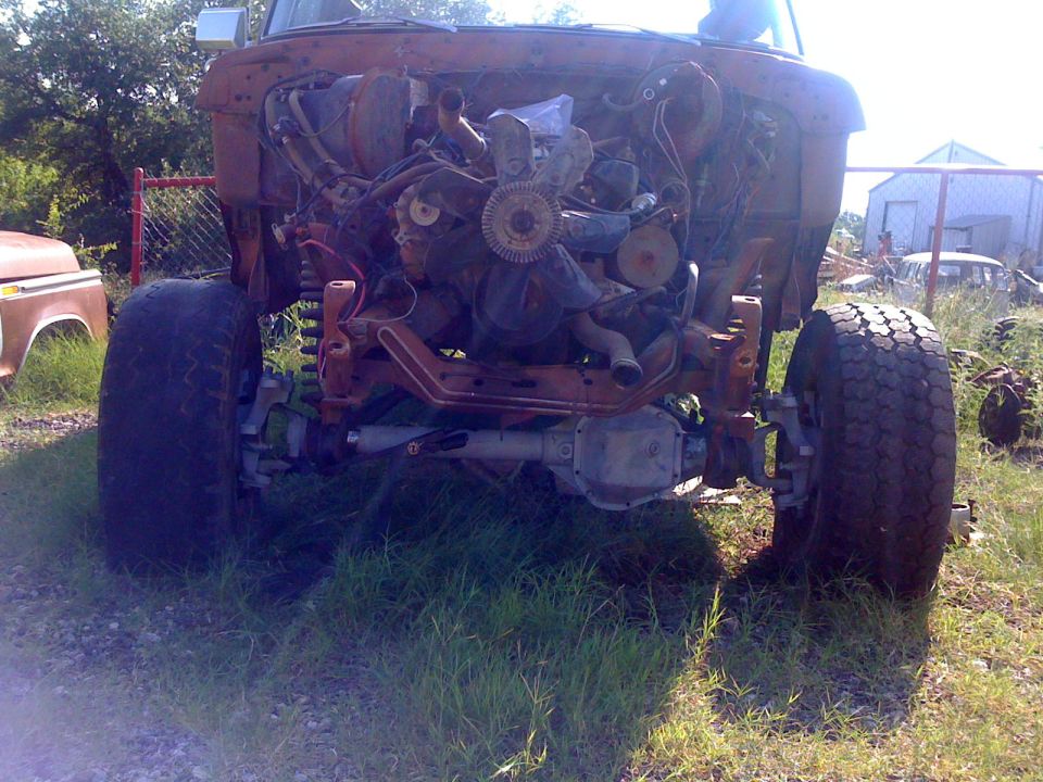 Front view of a rusty vehicle chassis with engine and suspension exposed. Another vehicle is partially visible to the left