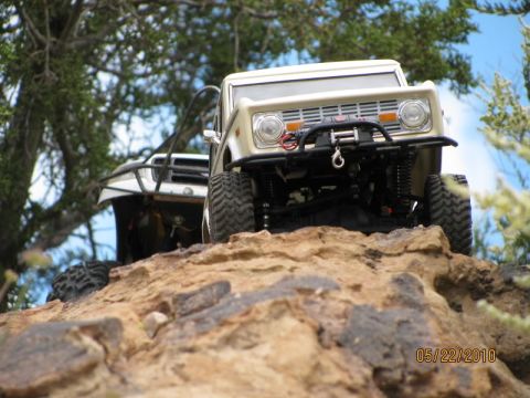 1988 Ford Bronco - Two remote-controlled trucks navigate a rocky terrain under a tree.