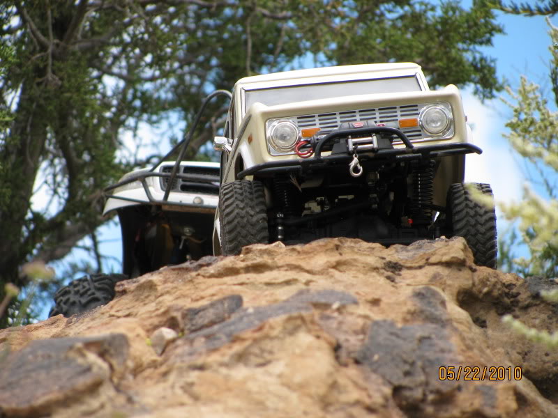 two toy trucks climbing rocks, one is tan and has a winch, the other is white