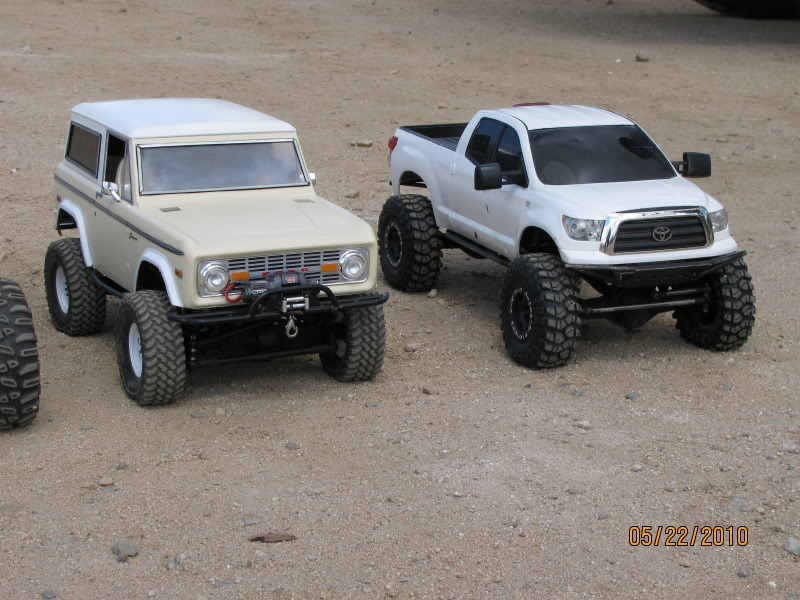 Two RC off-road vehicles, a cream-colored Bronco-style and a white Toyota Tundra-style, parked on dirt
