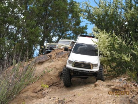 1988 Ford Bronco - Two remote control off-road vehicles navigate a challenging rocky incline under a blue sky.