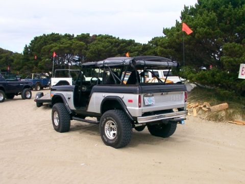 1980 Ford Bronco - This two-tone convertible SUV features a roll cage and is parked on a sandy beach with other vehicles in the background.