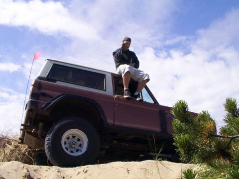 1980 Ford Bronco - A man sits atop a 1980 Ford Bronco, which is parked on a sandy incline under a cloudy sky.