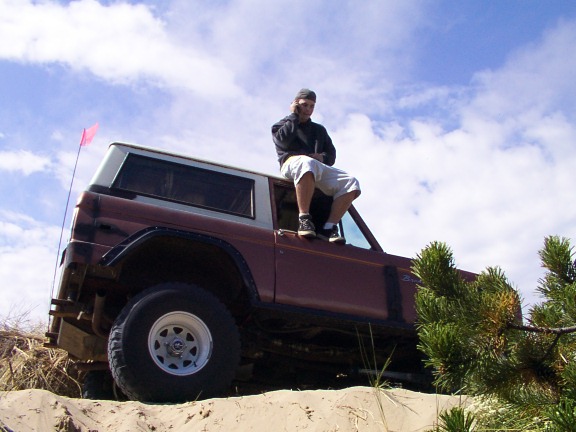 Maroon Ford Bronco SUV on sand dune with person sitting on hood and pink whip flag