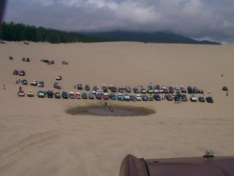 1980 Ford Bronco - A large gathering of off-road vehicles parked together on a sandy terrain under a cloudy sky.