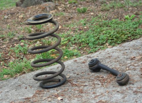 1976 Ford Ranchero - Two automotive suspension components, a rusty coil spring and a sway bar end link, are displayed on a concrete surface.