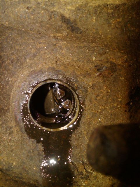 1982 Ford Bronco - The oil drain plug is visible in a rusty engine component, with dark oil pooling around it.