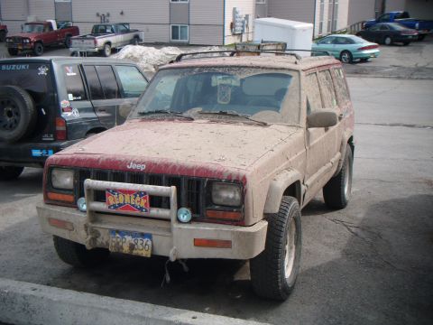 1998 Jeep Cherokee - Mud-covered red Jeep Cherokee parked next to another SUV, with a "Redneck" sticker on the grille guard.