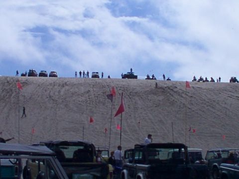 1980 Ford Bronco - A group of off-road vehicles and people are gathered on a large sand dune under a partly cloudy sky.