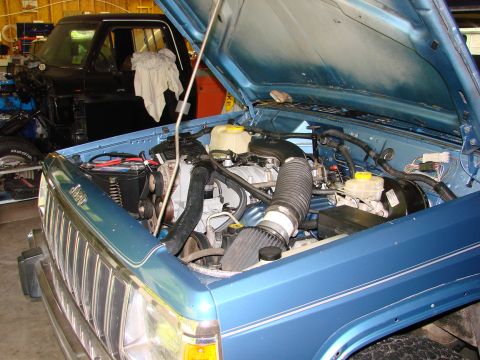 1991 Jeep Comanche - The engine bay of a blue Jeep Comanche is visible, with a black truck in the background.