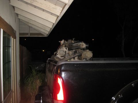 1969 Ford F-250 - An engine is loaded into the bed of a pickup truck at night, parked next to a house.