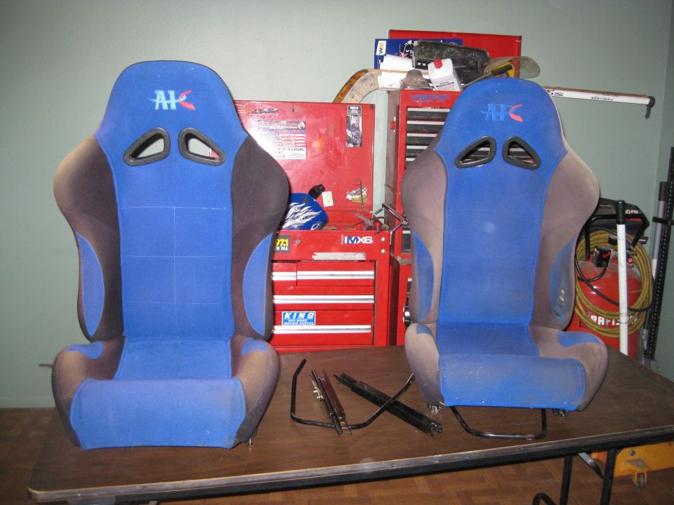 Pair of blue racing seats with AK logos, set on a workbench