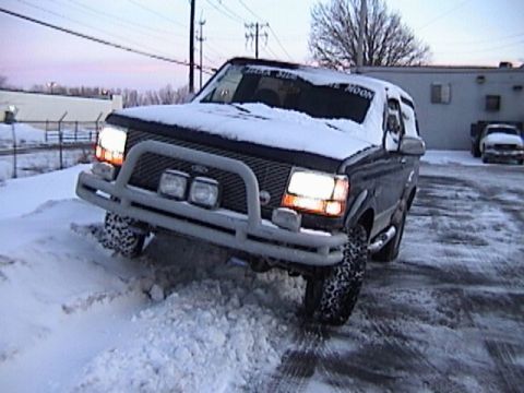 1995 Ford Bronco - A dark Ford Bronco navigates a snowy area with its headlights on.
