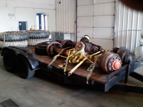 1985 Ford F-350 - A flatbed trailer carries multiple heavy-duty truck axles and spare tires, secured with yellow tie-down straps.