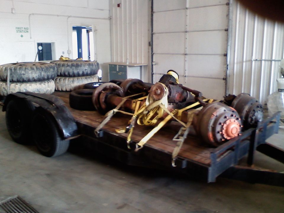 Trailer loaded with several large truck axles and spare tires. Some axles are strapped down with yellow straps