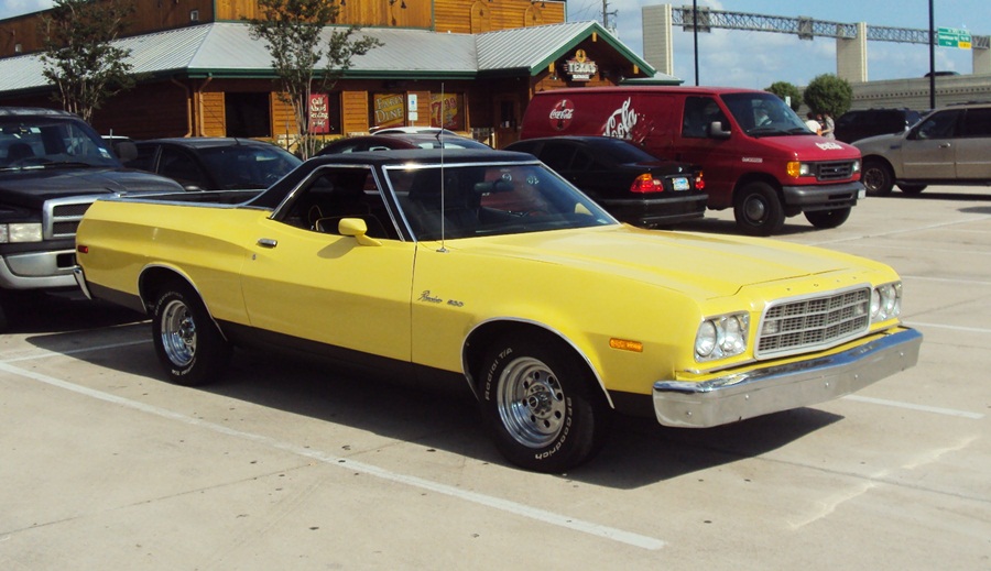 Yellow 1973 Ford Ranchero with black roof and chrome wheels parked in a lot near a log-style building