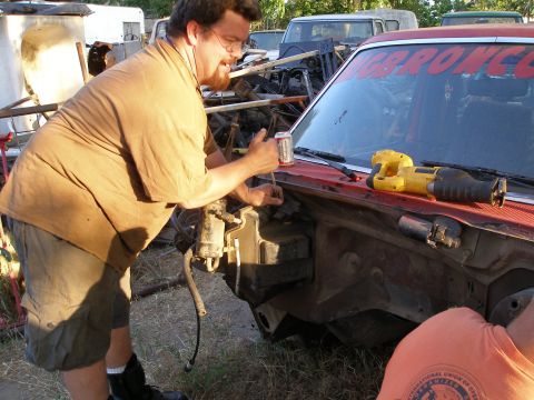 1969 Ford F-250 - Man works on the engine bay of a red vehicle, with a reciprocating saw nearby and 'BRONCO' text on the windshield.