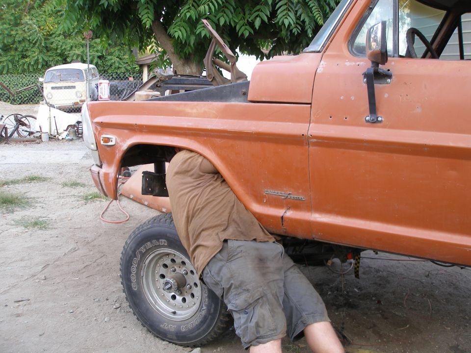 Orange pickup truck with a person underneath, side view. Visible text: SUPERMOTORS.NET