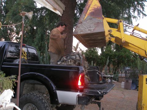 1969 Ford F-250 - A black pickup truck is positioned under a yellow loader bucket, with a man working near a tree.