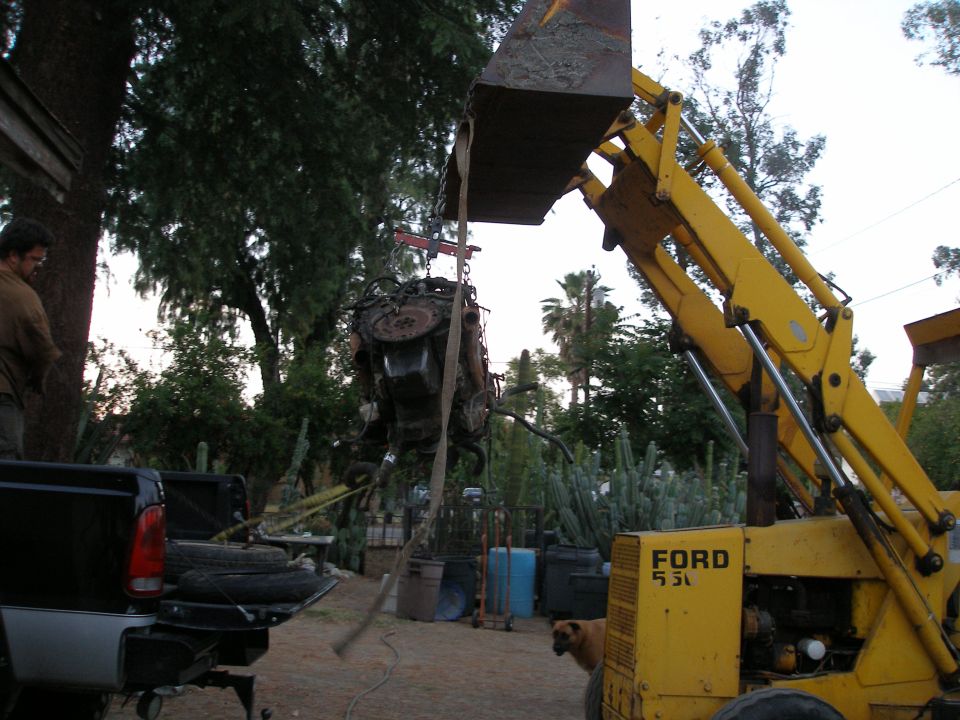 Engine being lifted by yellow backhoe; black truck bed in foreground with tires