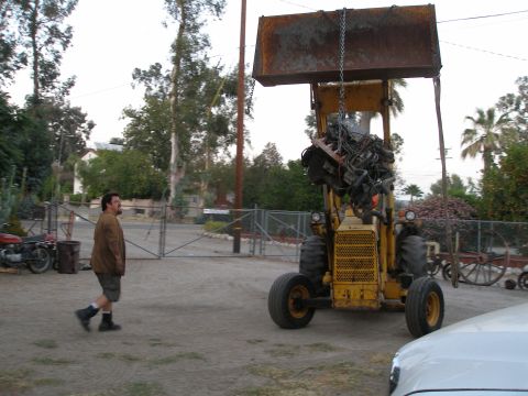 1969 Ford F-250 - A backhoe is lifting a complex engine assembly by a chain, with a man walking past in the foreground.