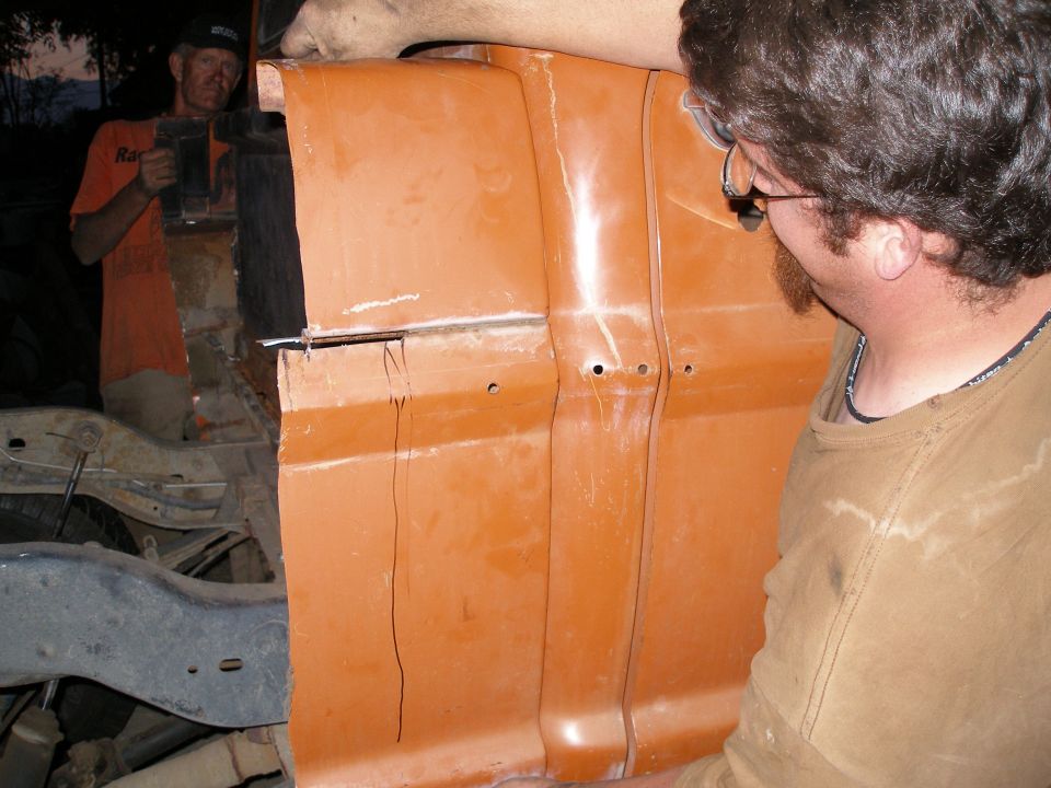 Orange truck cab section, viewed from the side, with two men working on it. The paint is faded