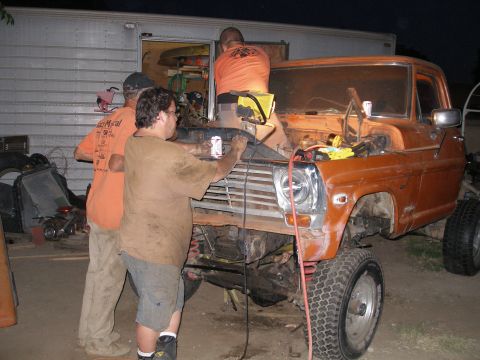 1969 Ford F-250 - Three men are gathered around the front of an orange pickup truck, appearing to be working on the engine.