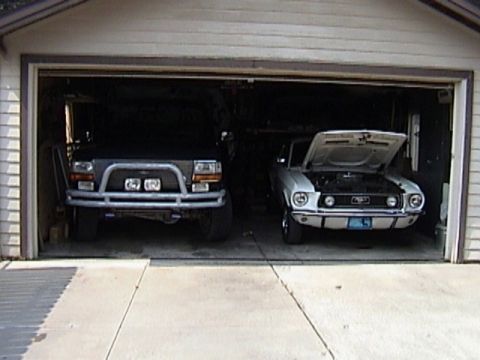 1995 Ford Bronco - Two vehicles are parked inside a garage, a black SUV and a white classic car with its hood up.