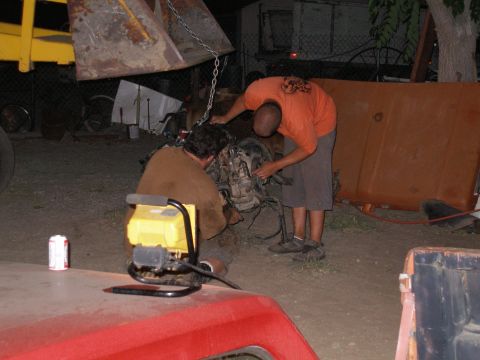1969 Ford F-250 - Two men are working on an engine that is suspended by chains from a yellow loader. A red truck is visible in the foreground.