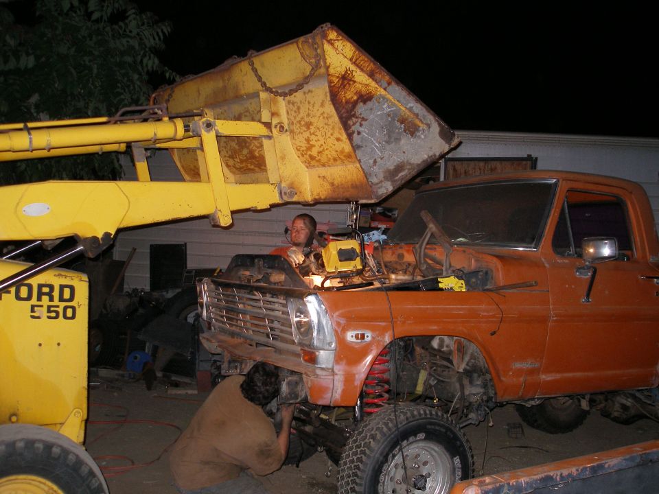 Orange Ford truck with hood open, two people working on it, front loader overhead. The truck appears to be a 1967-1972 Ford F-Series