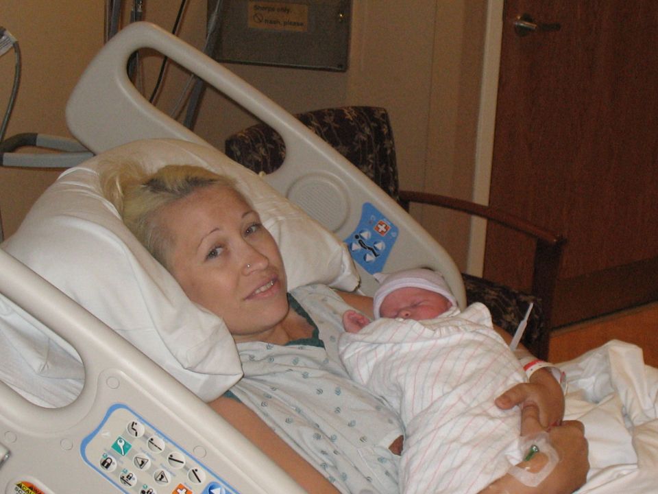 Woman in hospital gown holding a newborn baby in a hospital bed, with a "SUPERMOTORS.NET" watermark