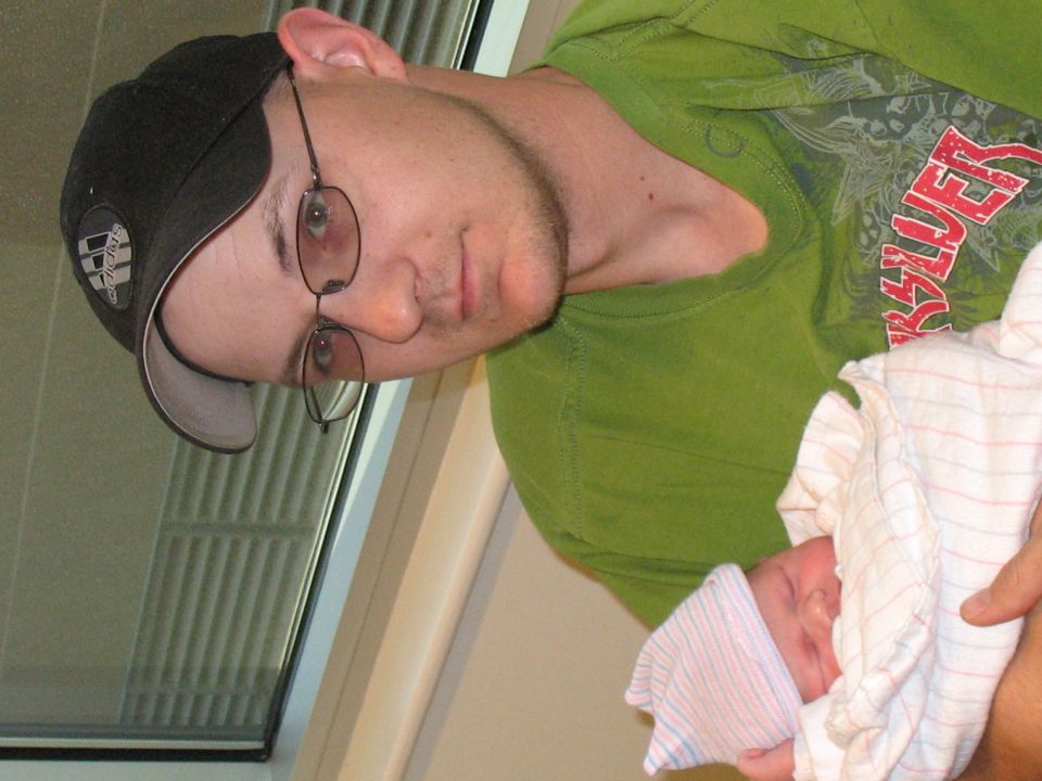 Man in a green shirt and black baseball cap holds a newborn baby wrapped in a blanket