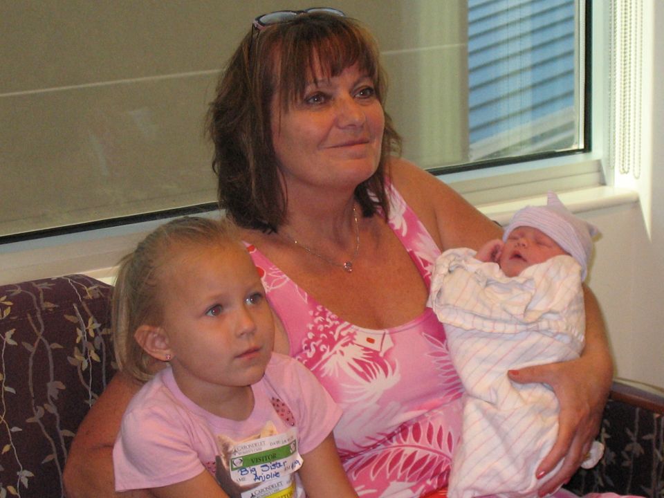 Woman in a pink floral dress holding a swaddled newborn and a young girl in a pink shirt