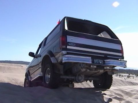 1995 Ford Bronco - A Ford Bronco navigates a sandy terrain, its rear profile against a bright sky.