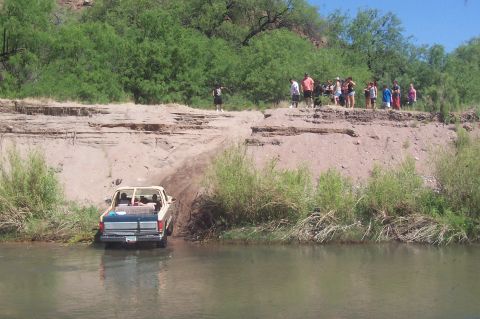 1981 Ford F-150 - A group of people watch a pickup truck enter the water near a sandy embankment.