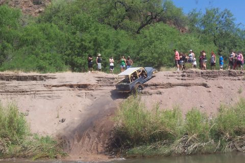 1981 Ford F-150 - A modified off-road vehicle navigates a steep dirt bank near a river, with spectators watching from above.