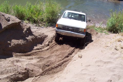 1981 Ford F-150 - A white Ford pickup truck navigates a challenging sandy and muddy terrain near a body of water.