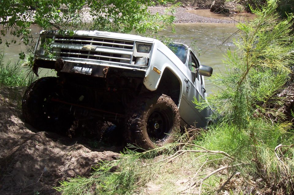 White square-body Chevy 4x4 climbing a muddy riverbank at a steep angle