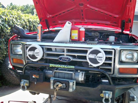 1985 Ford Bronco - Front view of a red Ford Bronco with its hood open, displaying maintenance supplies and a winch on the bumper.