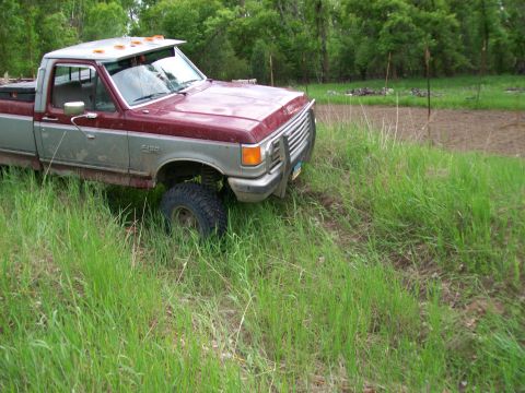 1988 Ford F-150 - A maroon and silver Ford F-150 pickup truck is partially obscured by tall grass, with a muddy area visible to the right.